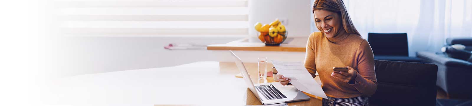 Photo of a smiling woman sitting at a table with an open laptop holding a piece of paper in one hand and her cell phone in the other.