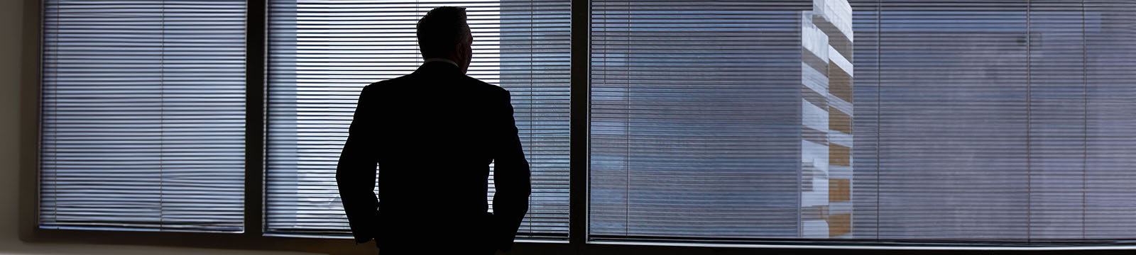 Photo of the silhouette of a man in suit looking out of a window to a cityscape.