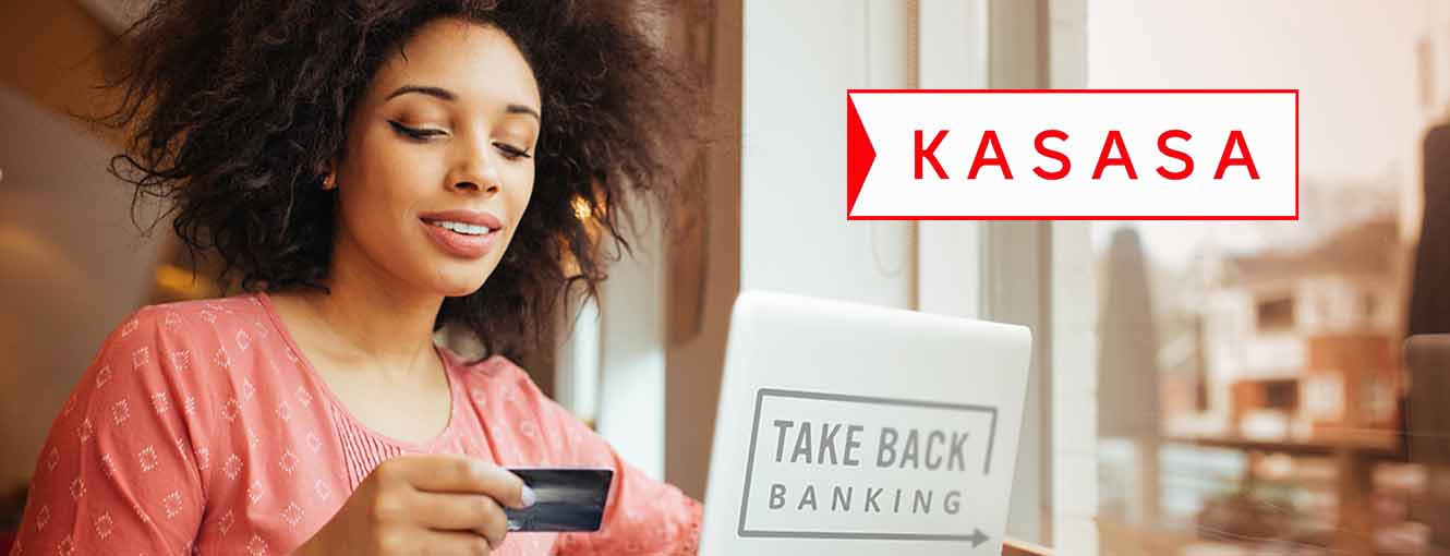 Woman holding a debit card while looking at her laptop. Laptop has a sticker on back that reads "Take Back Banking" and Kasasa logo is in upper right hand corner.