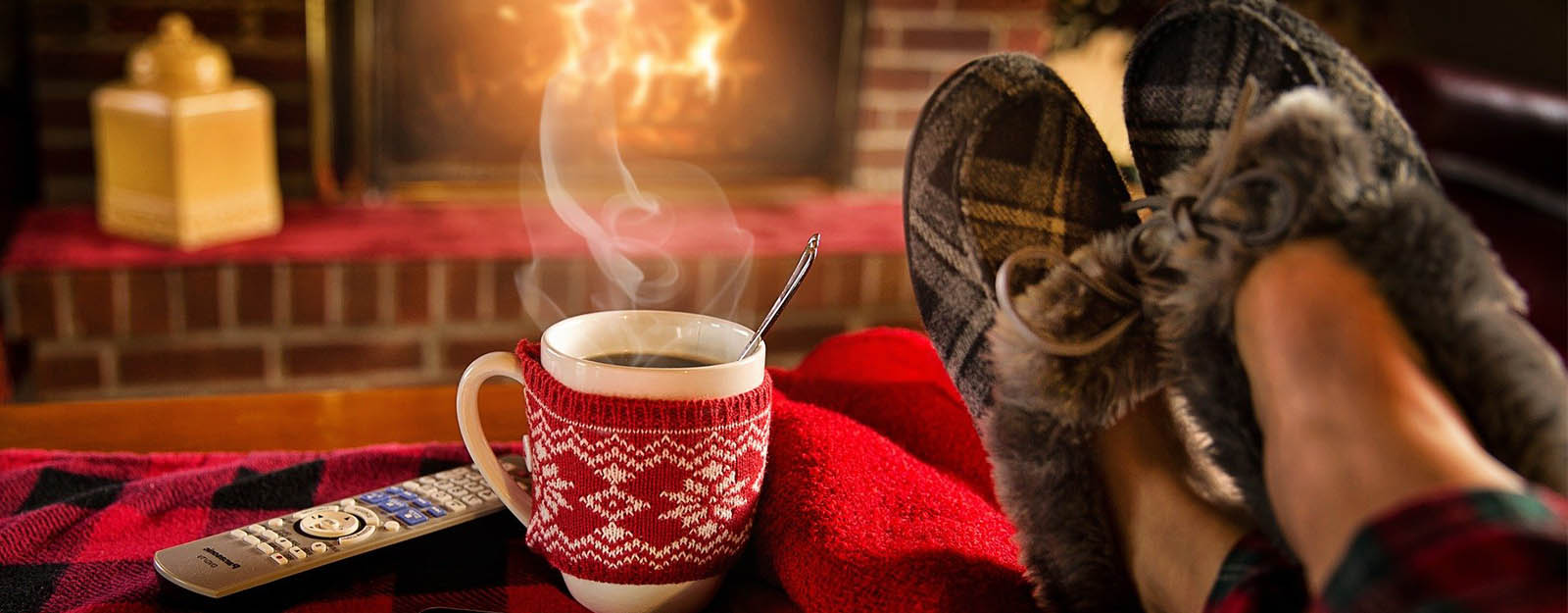 A pair of feet in slippers propped up beside a steaming mug of hot chocolate in front of a fire place.