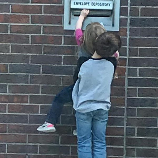 Photo of boy lifting up younger sister so that she can deposit an envelope in the night drop box.