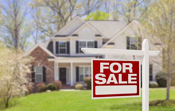 Photo of a for sale sign hanging in a yard with two story home in background.