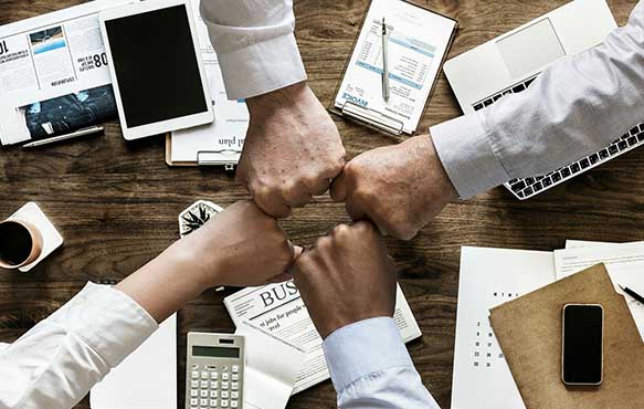 Photo of four individuals "fist bumping" across a table with various papers and office supplies underneath.