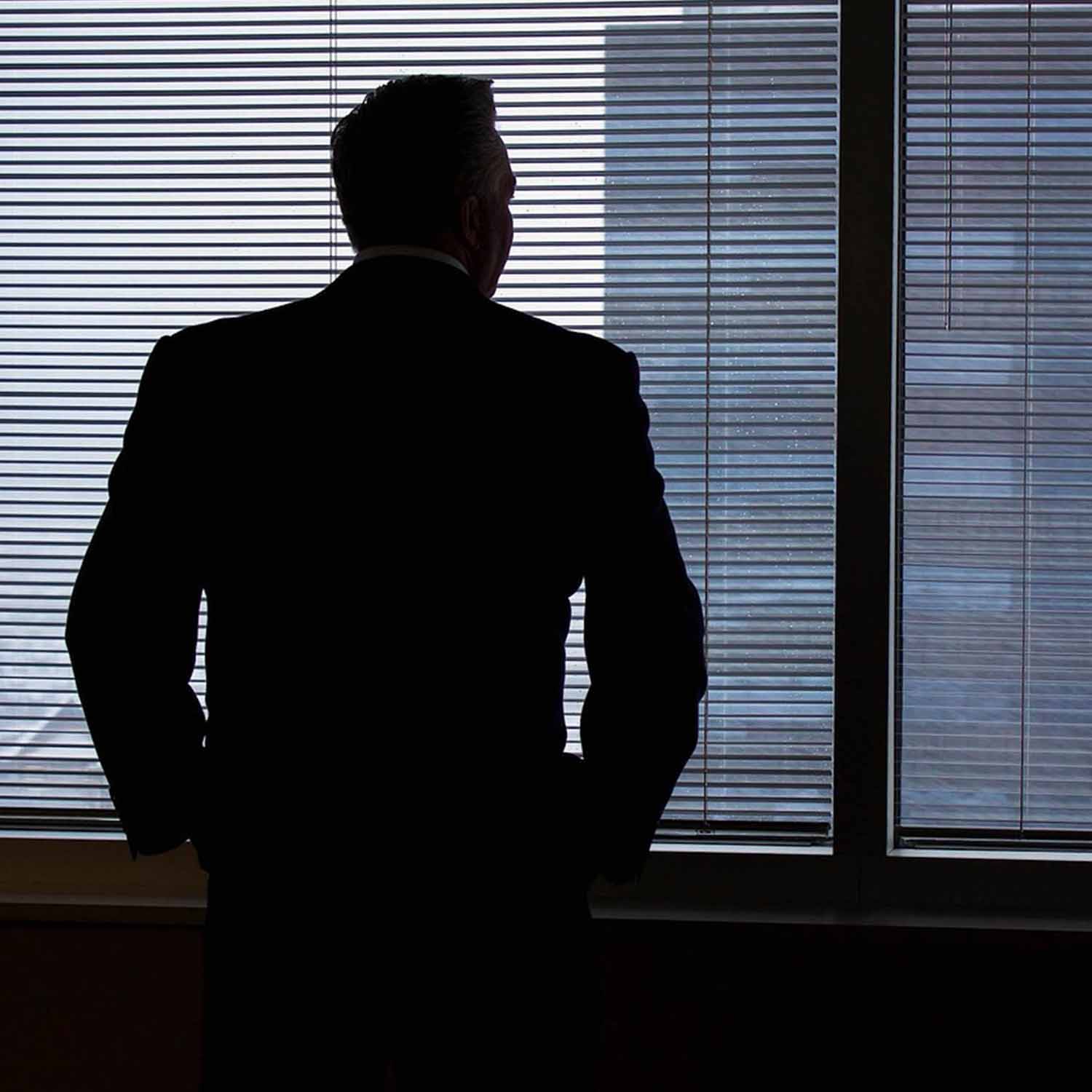 Photo of the silhouette of a man in suit looking out of a window to a cityscape.