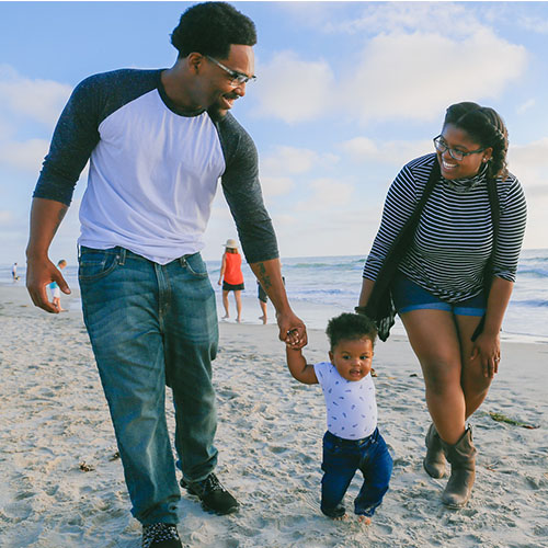 Young family holding the hands of a toddler while walking on the beach.