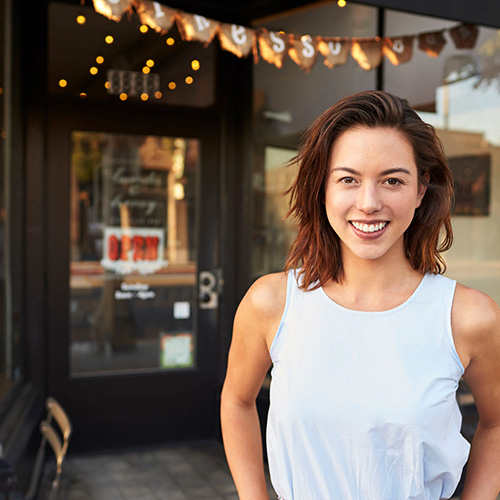 A business owner stands in front of a storefront with an open sign.